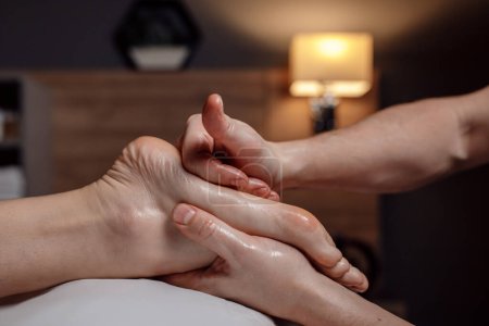 Relaxing foot massage, hands of a female massage therapist massaging female client foot in dark room of massage spa