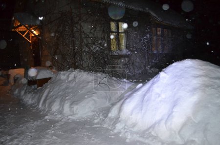 Foto de La fotografía muestra una casa salpicada de nieve en grandes cantidades, un camino despejado de nieve, un porche, iluminación eléctrica en las ventanas. - Imagen libre de derechos