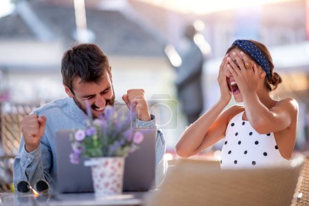 Foto de Imagen de pareja joven emocionada sentada en la cafetería, usando el ordenador portátil. - Imagen libre de derechos