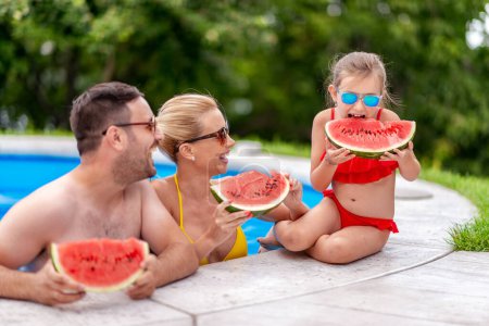 Téléchargez les photos : Famille assise au bord d'une piscine, s'amuser et manger de la pastèque. - en image libre de droit