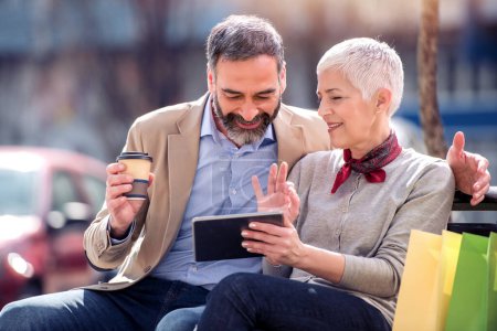 Téléchargez les photos : Couple âgé assis sur un banc dans le parc d'automne et en utilisant une tablette numérique
. - en image libre de droit