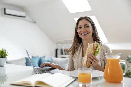 Photo for Smiling young woman having breakfast while working on laptop. - Royalty Free Image