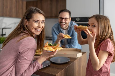 Mom, dad and daughter are eating together in kitchen.Theay are eating a big pizza.