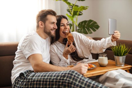 Téléchargez les photos : Jeune couple prenant le petit déjeuner dans le salon et prendre selfie avec tablette. - en image libre de droit