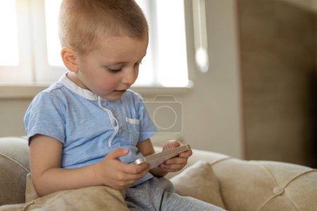 Foto de Lindo niño sentado en el sillón y jugando con el teléfono inteligente. - Imagen libre de derechos