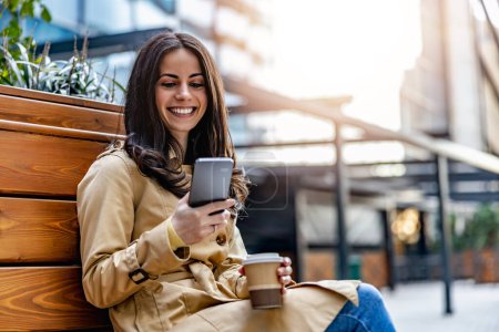 Foto de Mujer sentada en el banco en el parque de la ciudad, tomando café y usando teléfono móvil. - Imagen libre de derechos