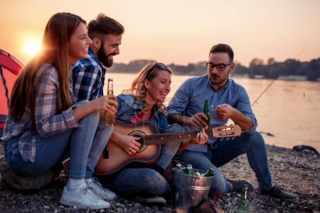 Téléchargez les photos : Groupe d'amis assis et écoutant la musique tout en buvant du cidre au coucher du soleil. - en image libre de droit