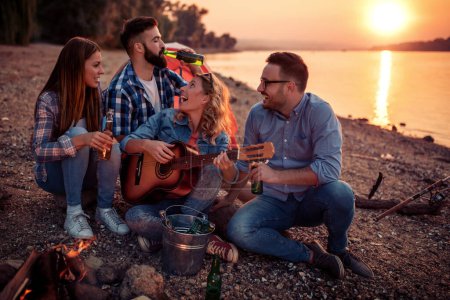 Photo for Group of young attractive friends are sitting next to river, playing the guitar and having fun together. - Royalty Free Image