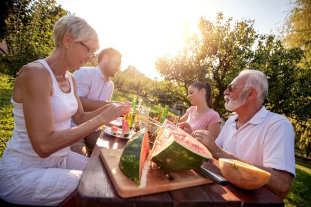 Foto de Dos generaciones de familia divirtiéndose mientras almuerzan en la naturaleza. Ocio, comida, gente y concepto de vacaciones. - Imagen libre de derechos
