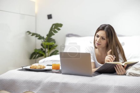 Foto de Retrato de una mujer joven haciendo notas en el portátil mientras está acostada en la cama en casa. - Imagen libre de derechos