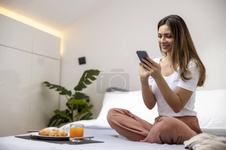 Photo for Woman having breakfast in bed while looking at mobile phone. - Royalty Free Image