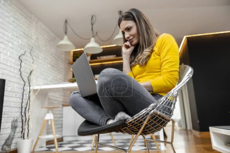 Photo for Beautiful young smiling woman working on laptop while sitting in comfortable chair at home. - Royalty Free Image