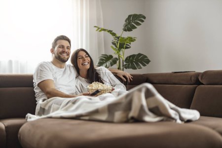 Photo for Young happy couple watching movie on TV and eating popcorn while relaxing in the living room. - Royalty Free Image