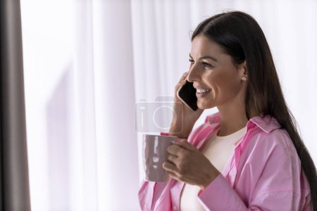Téléchargez les photos : Belle femme buvant du café et utilisant un smartphone à l'intérieur de sa maison à côté de la fenêtre lumineuse. - en image libre de droit