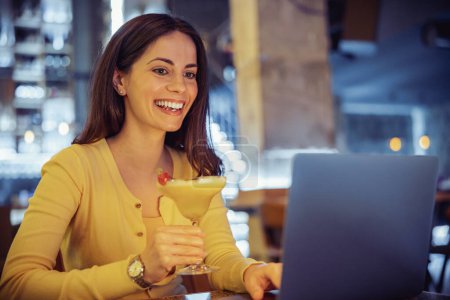 Foto de Hermosa chica bebiendo cóctel y sonriendo mientras trabaja en el ordenador portátil en cafe.Work desde la cafetería. - Imagen libre de derechos