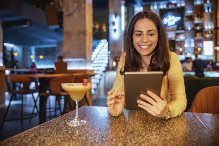 Téléchargez les photos : Jeune femme au bar prenant un cocktail et utilisant une tablette.Femme travaillant sur tablette dans un café moderne. - en image libre de droit