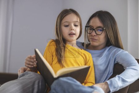 Photo for Happy mom and daughter smiling and reading book while relaxing on couch on weekend day at home. - Royalty Free Image