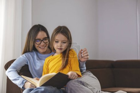 Photo for Happy mom and daughter smiling and reading book while relaxing on couch on weekend day at home. - Royalty Free Image