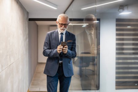Photo for Senior businessman using mobile phone in modern office.Confident senior director,entrepreneur, leader standing in his office. - Royalty Free Image