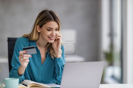 Téléchargez les photos : Jeune femme assise à une table dans le bureau avec ordinateur portable, travaillant à partir du bureau moderne. Achats en ligne. - en image libre de droit