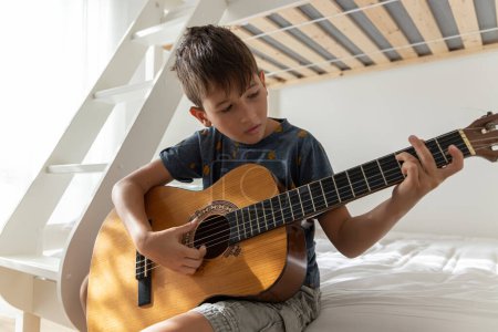 Téléchargez les photos : Il apprend à jouer de la guitare acoustique. Boy pratique la guitare acoustique dans sa chambre - en image libre de droit