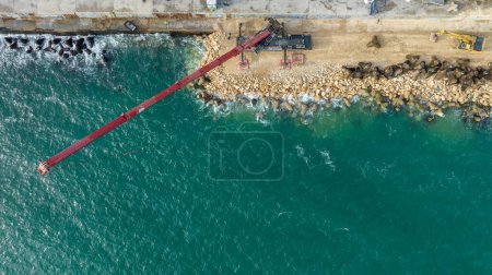 Foto de Vista aérea del sitio de construcción frente al mar con excavadora y grúa. Grúa trabajando en una construcción de rompeolas - Imagen libre de derechos