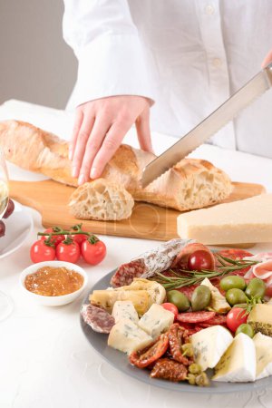 Photo for Woman serving traditional Italian antipasti and cutting a baguette on white table. Charcuterie plate with different types of sausages and cheeses - salami, dorblu, proscuitto served with olives and tomatoes - Royalty Free Image
