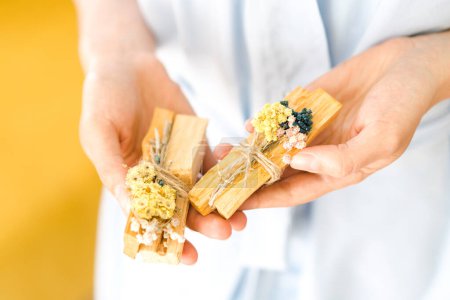 Photo for Woman hands holding palo santo sticks decorated with dry flowers and fragrant herbs against yellow wall. Fumigation ritual process. Burning palo santo for meditation - Royalty Free Image