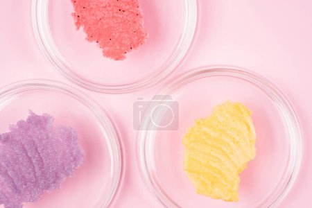 Téléchargez les photos : Sugar body scrub smears texture. Pink red, purple and yellow scrub smudges in petri glass dishes over pastel pink background. Skin care product with fruit extract - en image libre de droit