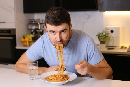Téléchargez les photos : Portrait de beau jeune homme mangeant des spaghettis à la cuisine - en image libre de droit