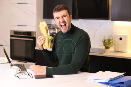 Téléchargez les photos : Portrait de beau jeune homme mangeant un sandwich tout en travaillant de la maison à la cuisine - en image libre de droit