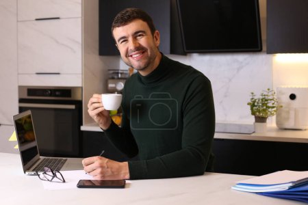 Foto de Retrato de joven guapo con taza de café trabajando desde casa en la cocina - Imagen libre de derechos