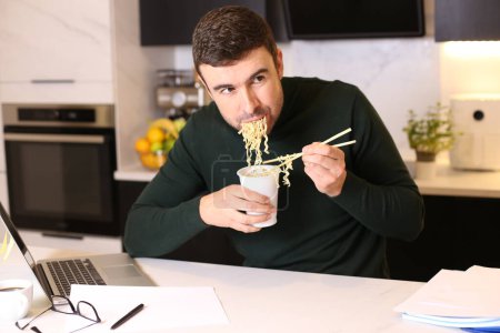 Foto de Retrato de joven guapo comiendo fideos taza mientras trabaja desde casa en la cocina - Imagen libre de derechos
