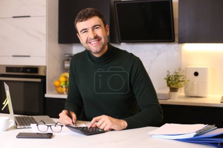 Téléchargez les photos : Portrait de beau jeune homme travaillant de la maison avec ordinateur portable à la cuisine - en image libre de droit