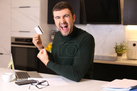 Foto de Retrato de joven guapo haciendo e-shopping con tarjeta de crédito y portátil en la cocina - Imagen libre de derechos