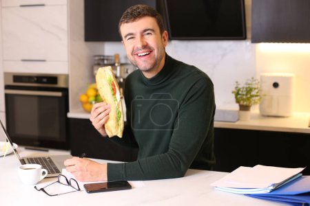 Foto de Retrato de joven guapo comiendo sándwich mientras trabaja desde casa en la cocina - Imagen libre de derechos