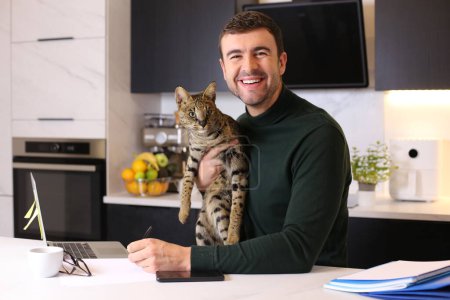 Téléchargez les photos : Portrait de beau jeune homme avec son chat exotique assis à la cuisine - en image libre de droit