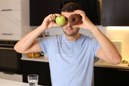 Foto de Retrato de un joven guapo sosteniendo una manzana sana y un donut poco saludable frente a sus ojos en la cocina - Imagen libre de derechos