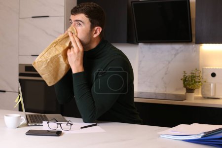 Foto de Retrato de joven guapo respirando con bolsa de papel mientras trabaja desde casa en la cocina - Imagen libre de derechos