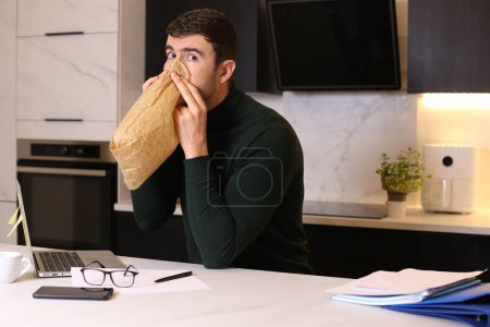 Téléchargez les photos : Portrait de beau jeune homme respirant avec un sac en papier tout en travaillant à la maison à la cuisine - en image libre de droit