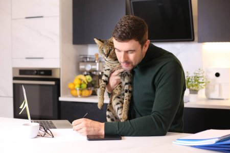 Téléchargez les photos : Portrait de beau jeune homme avec son chat exotique assis à la cuisine - en image libre de droit