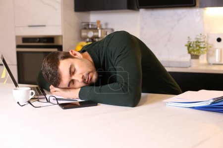 Foto de Retrato de joven guapo durmiendo en la mesa mientras trabaja desde casa en la cocina - Imagen libre de derechos