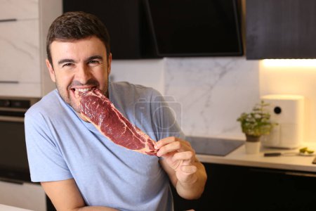 Téléchargez les photos : Portrait de beau jeune homme tenant un steak de viande crue à la cuisine - en image libre de droit