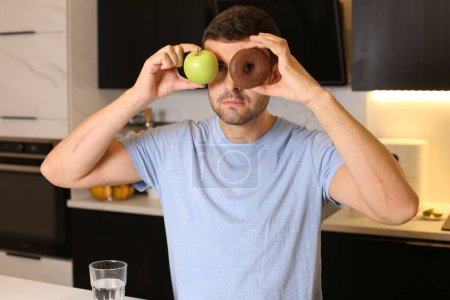 Foto de Retrato de un joven guapo sosteniendo una manzana sana y un donut poco saludable frente a sus ojos en la cocina - Imagen libre de derechos