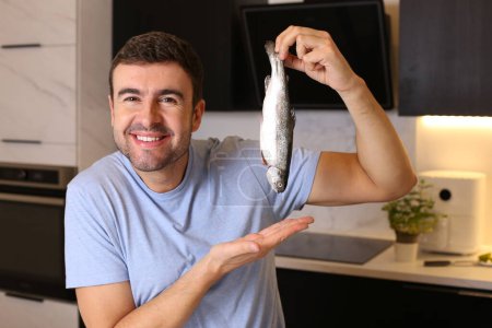 Téléchargez les photos : Portrait de beau jeune homme avec du poisson cru à la cuisine - en image libre de droit