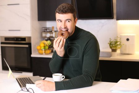 Foto de Retrato de joven guapo comiendo donut con café mientras trabaja desde casa en la cocina - Imagen libre de derechos
