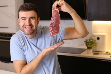 Photo for Portrait of handsome young man holding raw meat steak at kitchen - Royalty Free Image