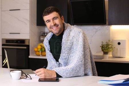 Téléchargez les photos : Portrait de beau jeune homme malade avec couverture travaillant de la maison à la cuisine - en image libre de droit