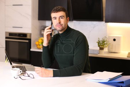Foto de Retrato de joven guapo hablando por teléfono mientras trabajaba desde casa en la cocina - Imagen libre de derechos