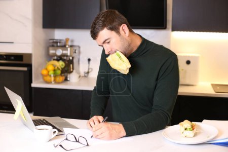 Photo for Portrait of handsome young man eating sandwich while working from home at kitchen - Royalty Free Image
